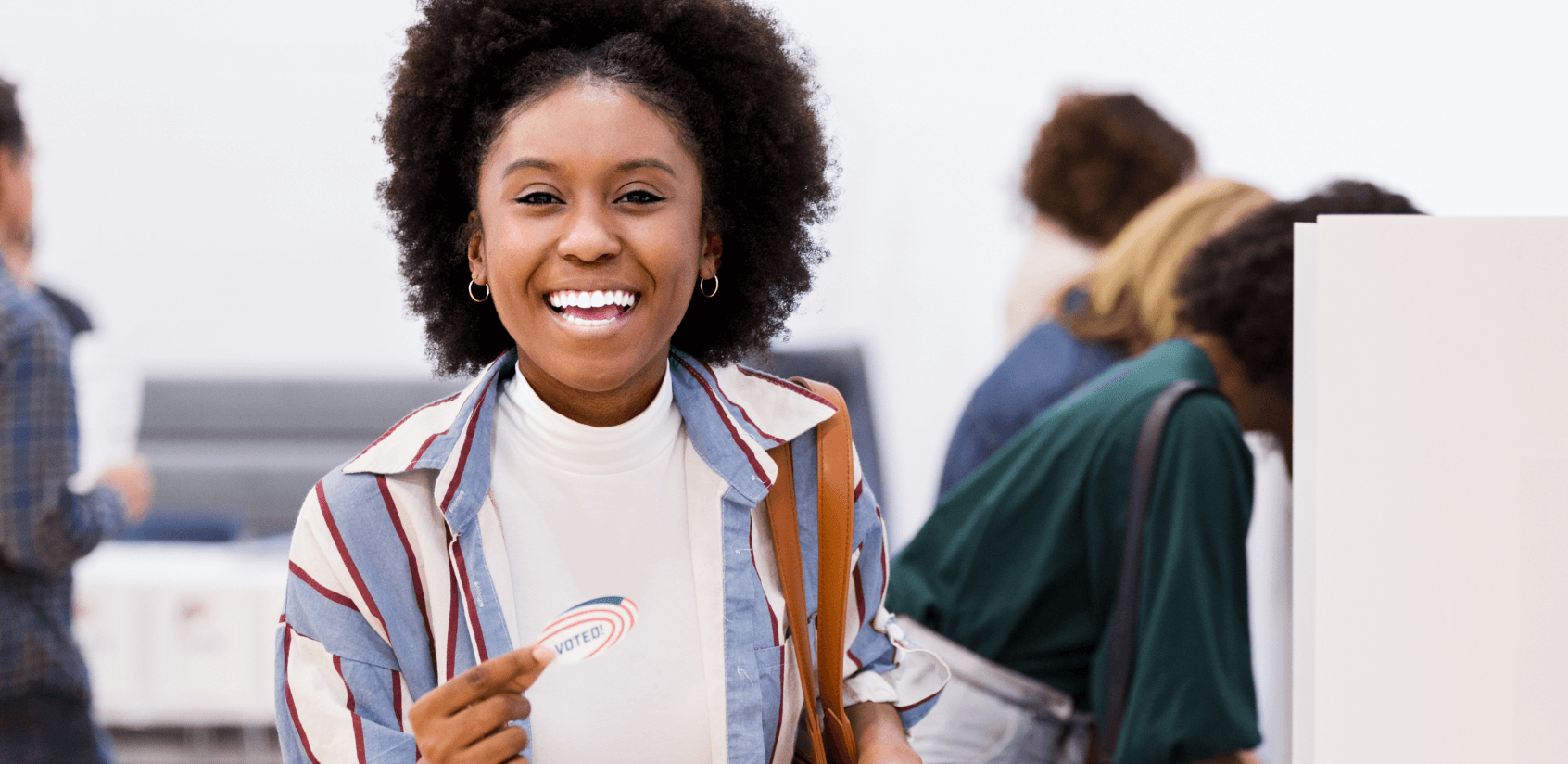 Woman standing next to a voting booth smiling and holding an “I voted” sticker.