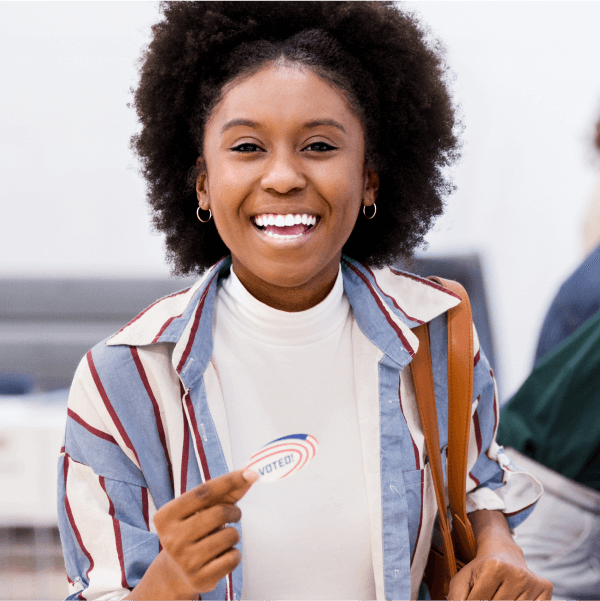 Woman standing next to a voting booth smiling and holding an “I voted” sticker.