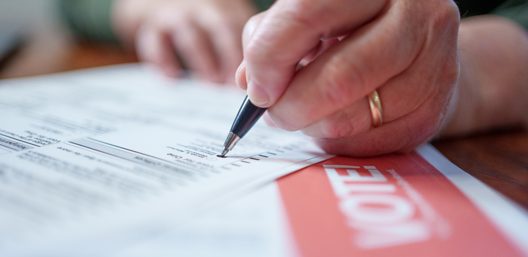 Close-up image of a hand holding a pen while filling out a mail-in ballot.